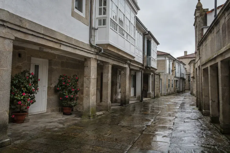 Calle peatonal con soportales de granito, galerías blancas de madera y el suelo de losa mojado