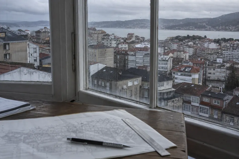 Mesa con planos ante el tejido urbano en ladera y la ría de Vigo bajo un cielo de lluvia